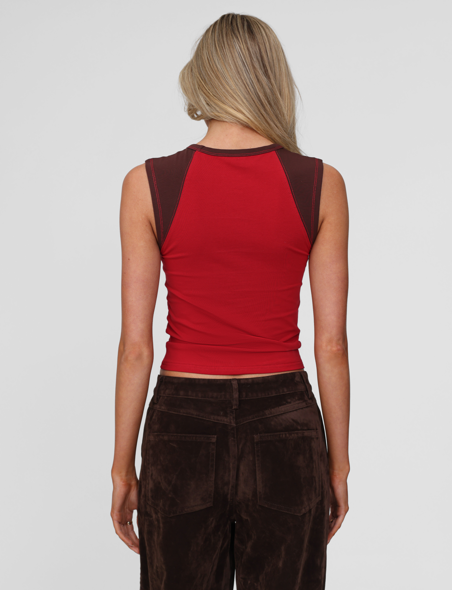 A woman with long blonde hair is shown from behind, wearing the TIGERMIST FIERA TEE in red. She pairs it with dark brown velvet pants against a plain, light-colored background.