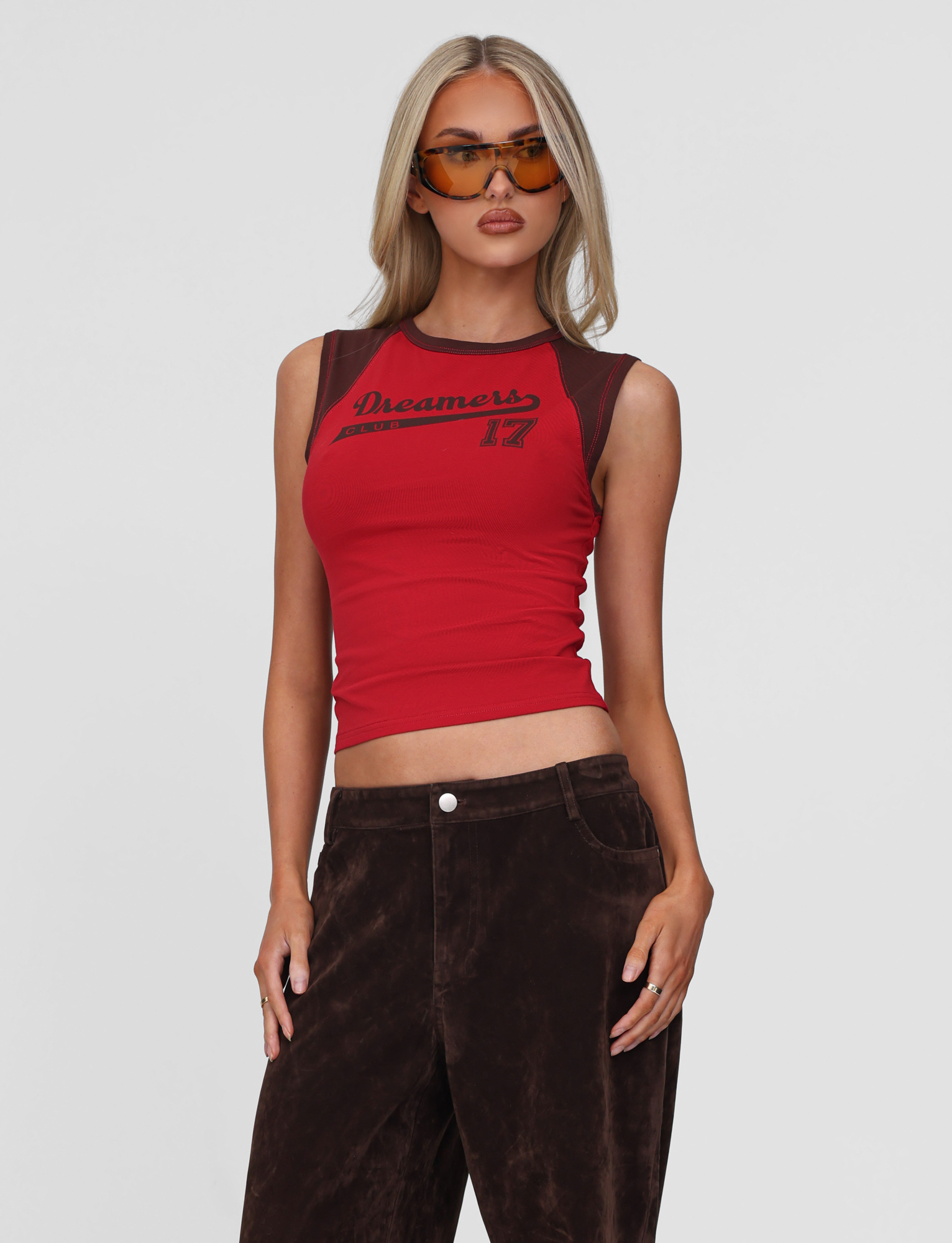 A woman with long blonde hair and large brown sunglasses wears the TIGERMIST FIERA TEE - RED in an exclusive print, paired with dark brown velvet pants, posing with one hand in her pocket against a plain light background.