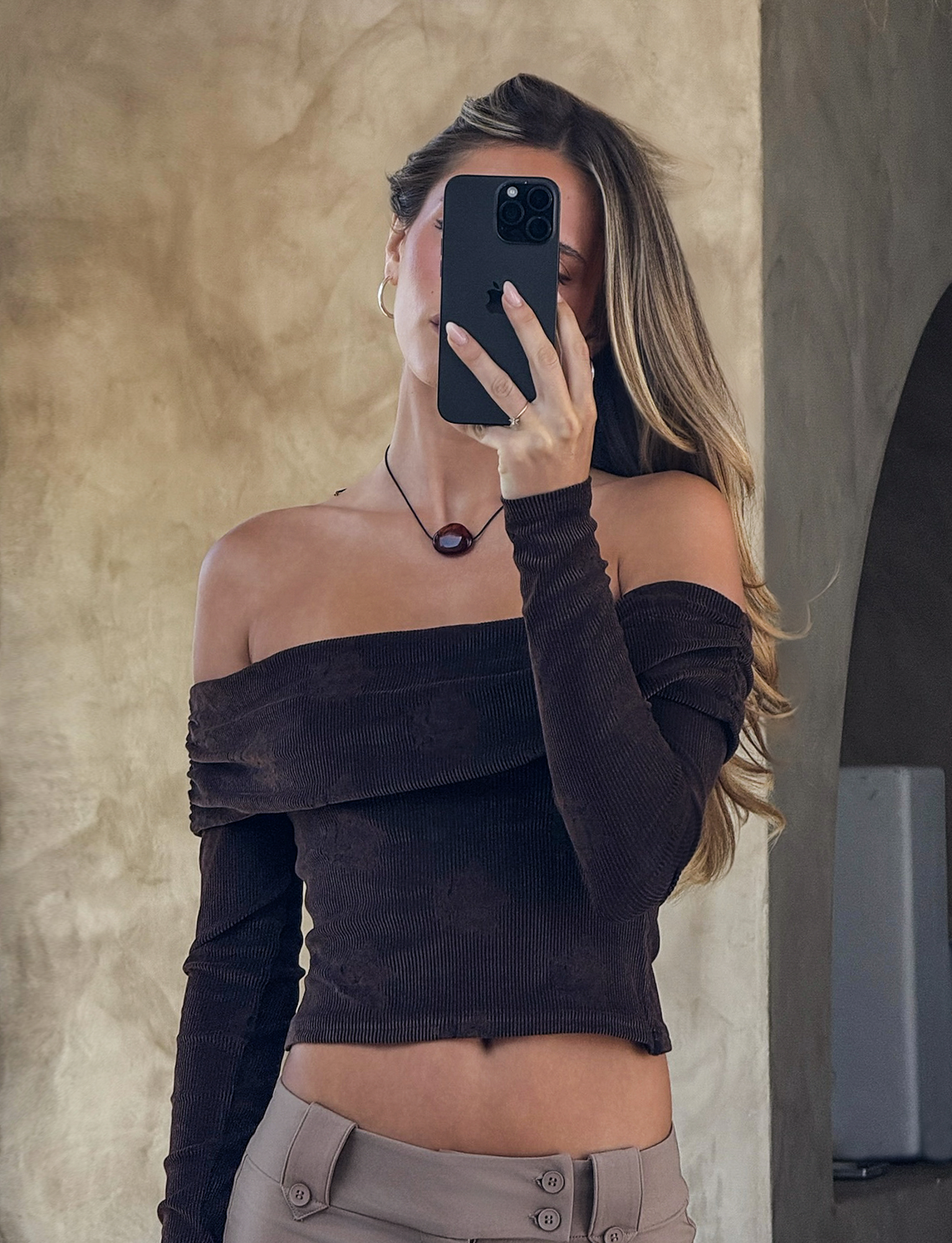 A woman with long, wavy hair snaps a mirror selfie, covering her face with her phone. She wears the TIGERMIST KORRA TOP in Chocolate Rose, paired with beige pants and a round necklace against a neutral textured wall.