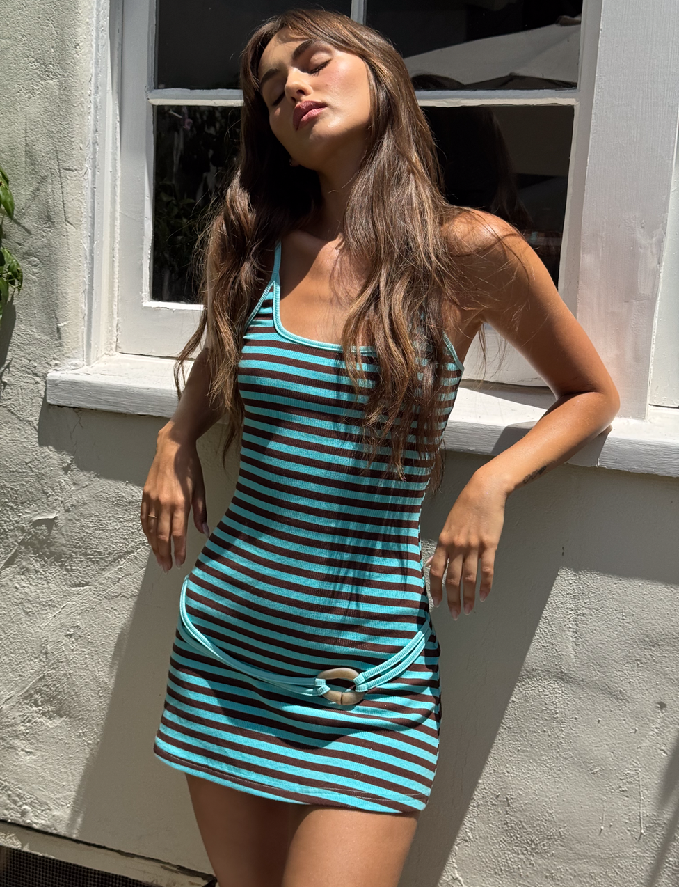 A woman stands outside in the TIGERMIST CELESTE MINI DRESS - BLUE STRIPE, eyes closed and facing the sun. Her long brown hair flows as she knots the hem of her soft ribbed dress in front of a white window and wall.
