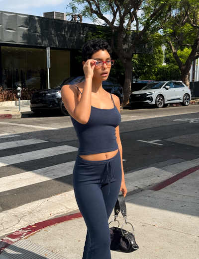 A person wearing the TIGERMIST LEORA TOP in navy, paired with matching pants, stands on a city sidewalk holding sunglasses and a small black handbag, with trees and cars visible in the background.