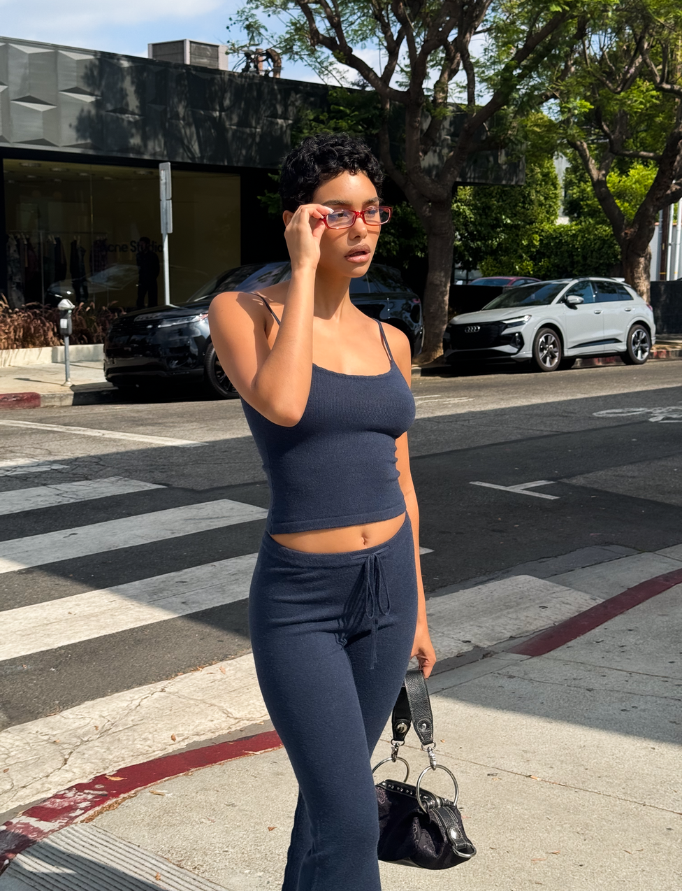 A person wearing the TIGERMIST LEORA TOP in navy, paired with matching pants, stands on a city sidewalk holding sunglasses and a small black handbag, with trees and cars visible in the background.