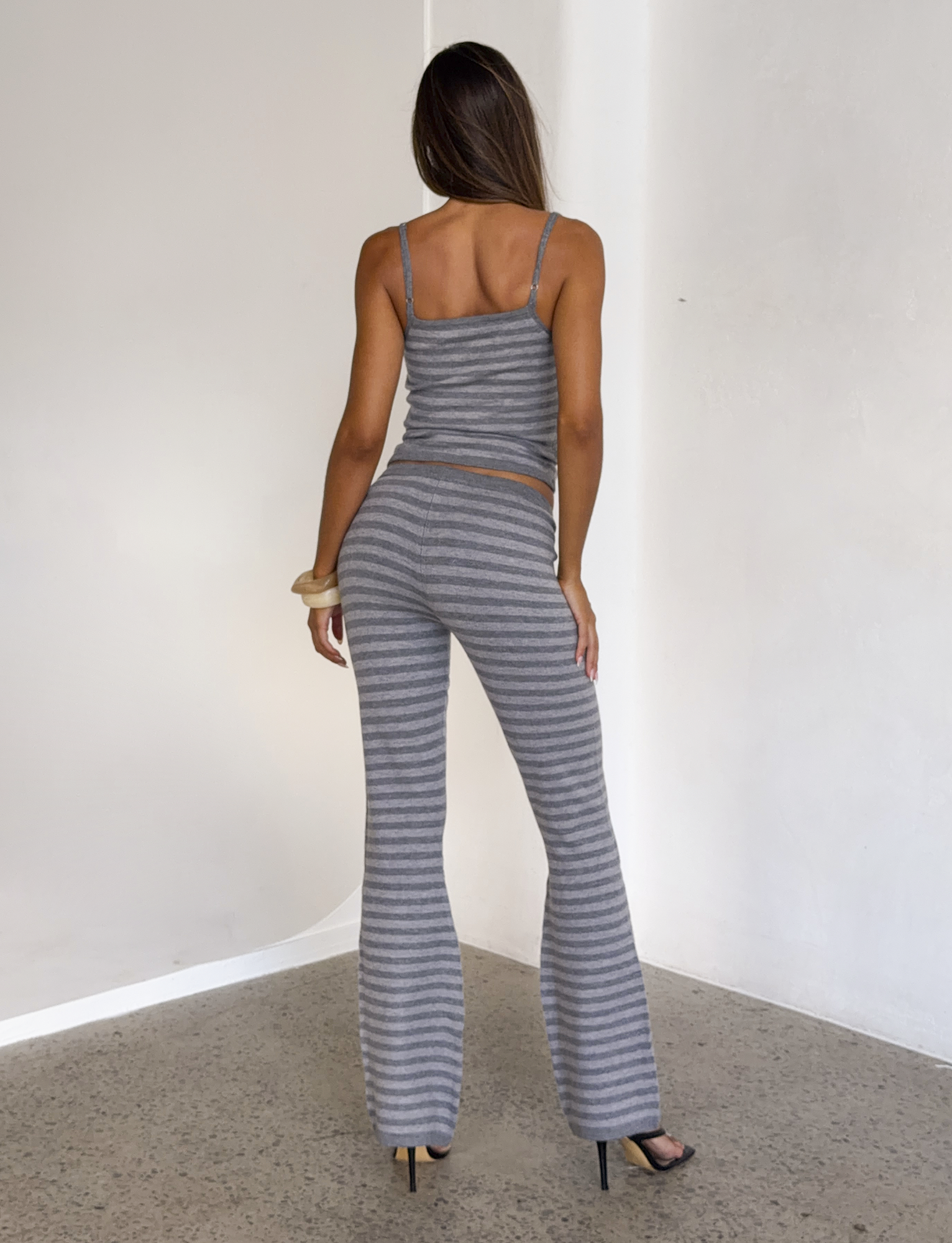 A woman with long hair, wearing high heels, models the TIGERMIST LEORA PANT - GREY STRIPE with a drawstring waist and a matching knit top indoors against light walls and a speckled floor, facing away.