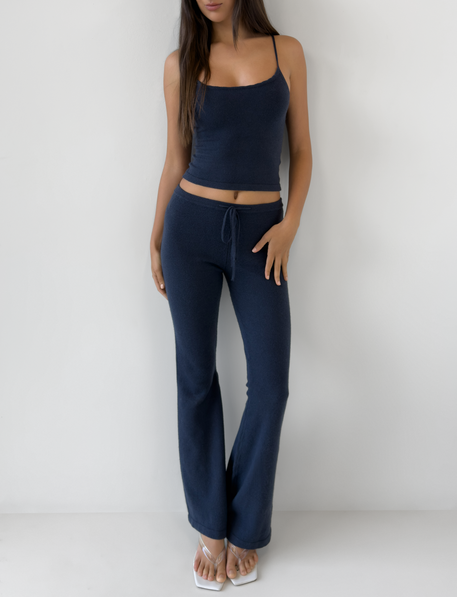 A woman stands against a white wall wearing the TIGERMIST LEORA PANT in navy—relaxed fit with a drawstring waist and flared legs—styled with a navy blue sleeveless crop top, white flip-flops, and her long hair down, one hand on her hip.