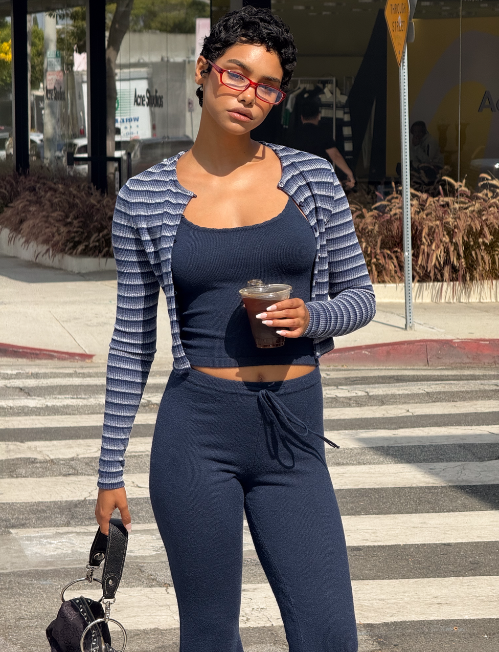 A woman in a navy two-piece set stands on a crosswalk holding an iced coffee and small bag. She wears the TIGERMIST TIMARA CARDIGAN in NAVY STRIP, red glasses, and has short hair. Buildings and plants decorate the background.