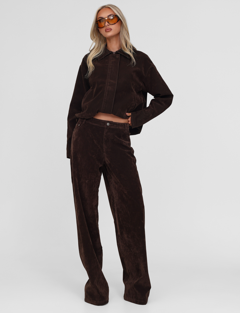 A woman poses confidently against a white background in TIGERMIST's CAMELLIA PANT—chocolate suede, styled with a loose, dark brown jacket and orange-tinted sunglasses. She has long blonde hair.