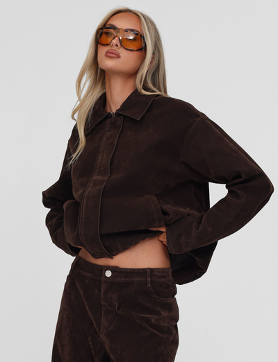 A woman with long blonde hair models the TIGERMIST CAMELLIA BOMBER JACKET in chocolate suede, pairing it with matching pants and large amber sunglasses. She stands confidently with one hand in her pocket against a light background.