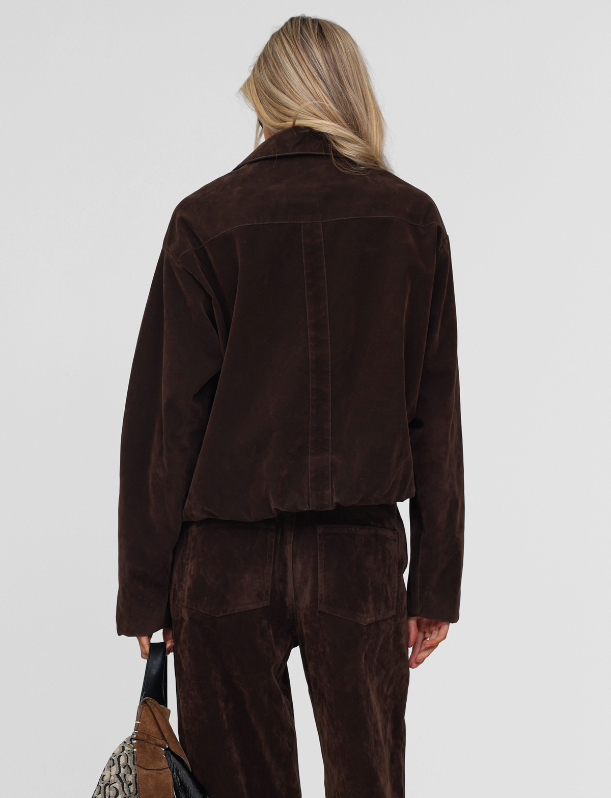 Seen from behind, a person with long blonde hair wears the TIGERMIST CAMELLIA BOMBER JACKET in chocolate suede and matching pants, holding a snakeskin-pattern faux leather bag. The backdrop is plain and light-colored.