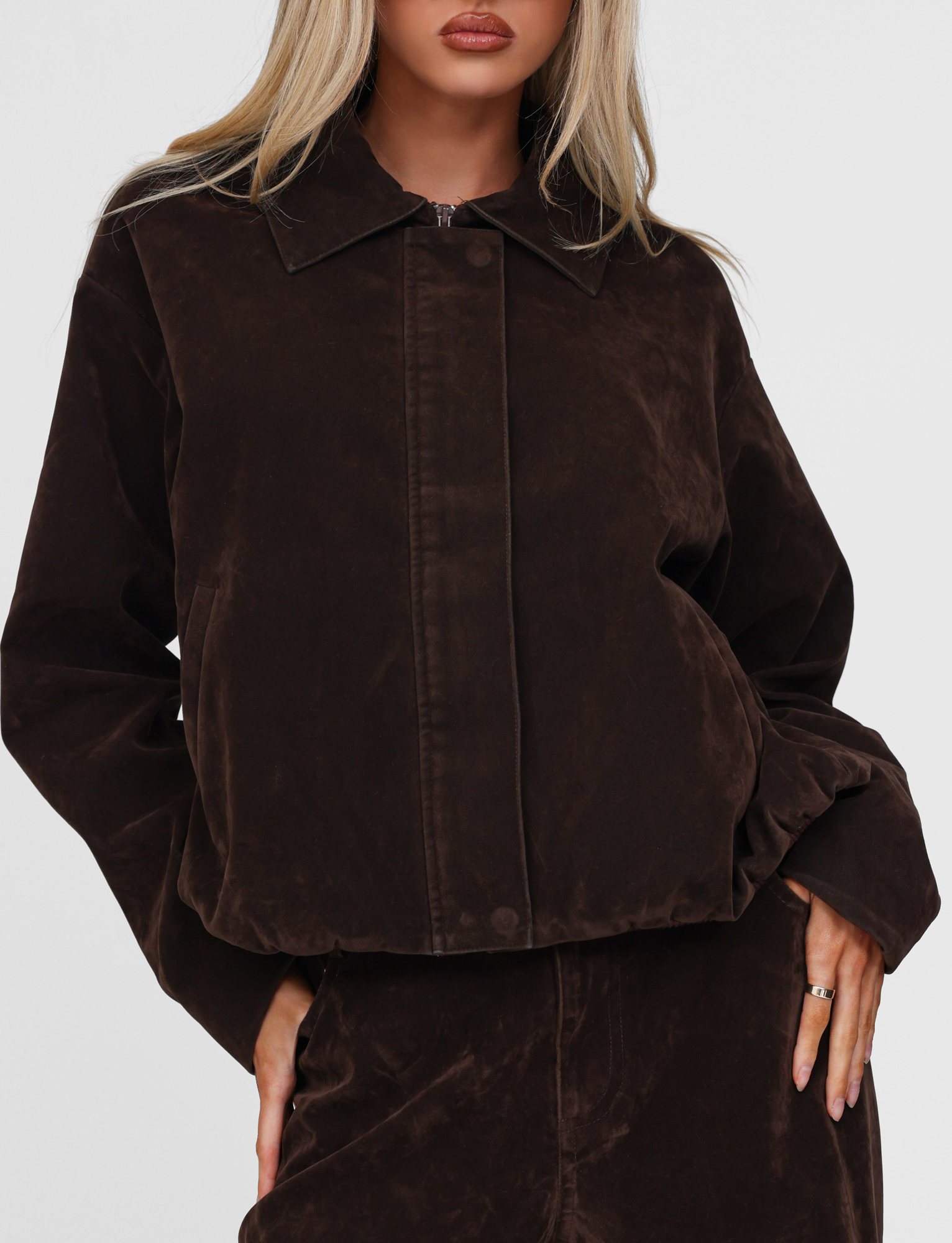 A woman models the TIGERMIST CAMELLIA BOMBER JACKET in chocolate suede, paired with a matching skirt. She stands against a plain white background, her blonde hair loose and face partially visible, one hand resting on her hip.