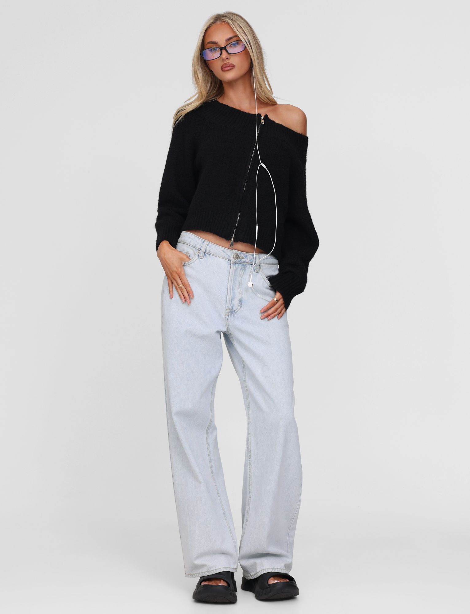 A woman with long blonde hair poses confidently against a plain background wearing TIGERMIST's LUNETTE JUMPER in black, paired with purple glasses, light blue wide-leg jeans, black sandals, and earphones around her neck.