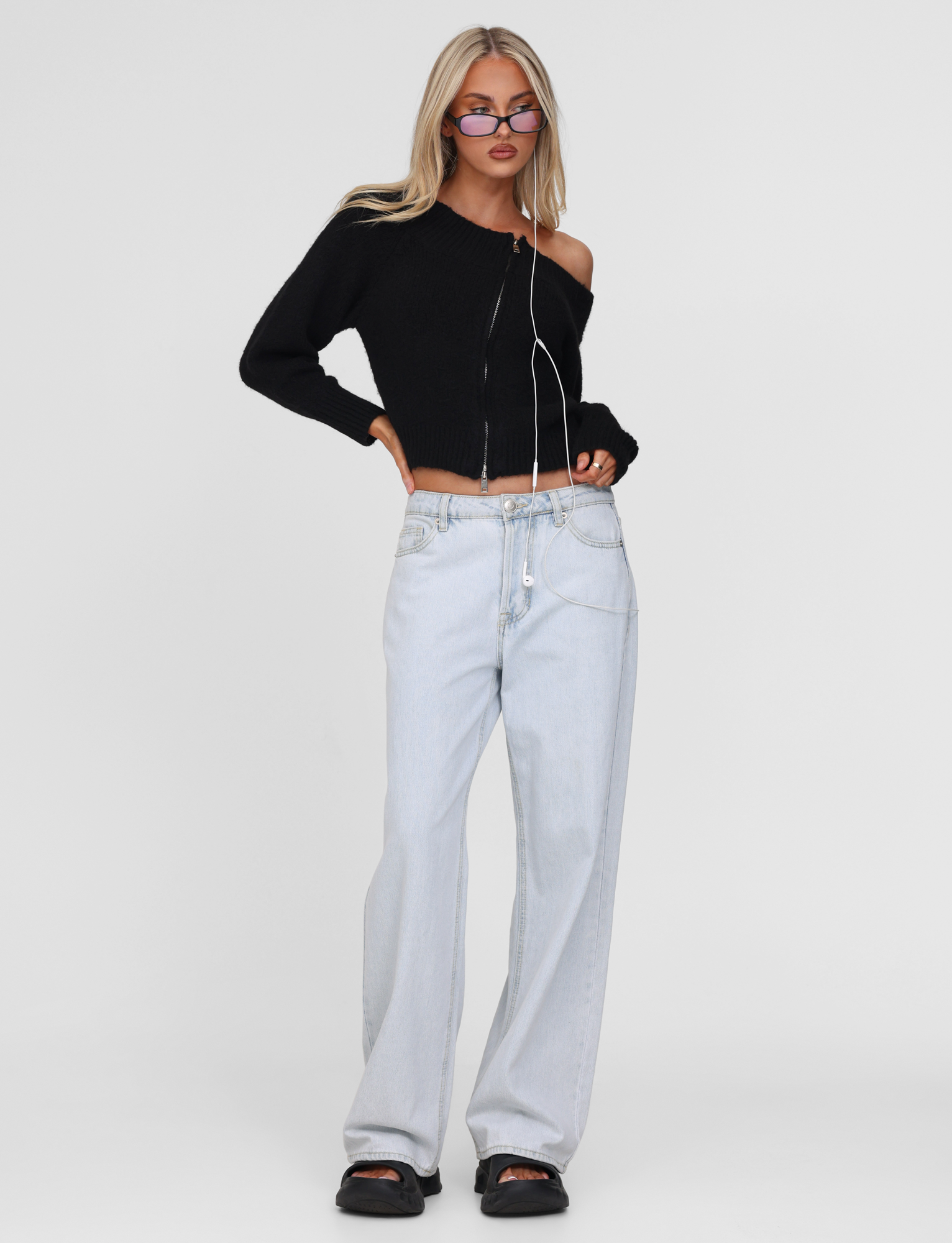 A woman poses against a plain background wearing the TIGERMIST LUNETTE JUMPER in black, paired with light-wash baggy jeans, black sandals, sunglasses, and a long necklace, her hand tucked in her pocket.