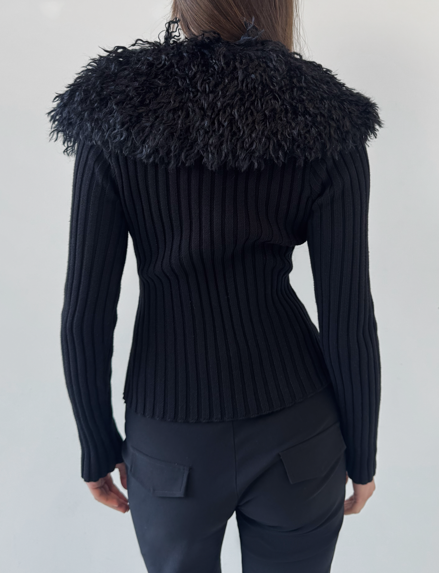 A woman is seen from the back wearing the TIGERMIST BRISA JUMPER in black, featuring a ribbed knit and textured faux fur collar, paired with black pants with back pockets, set against a plain light background.