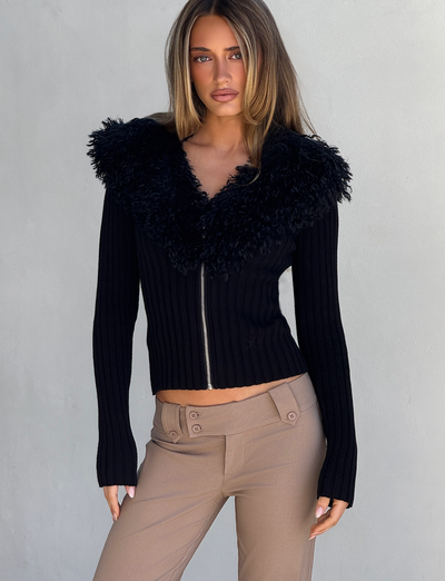 A woman with straight, light brown hair models the TIGERMIST BRISA JUMPER in black, featuring a ribbed texture and faux fur collar. She pairs it with taupe pants and stands against a plain light gray background, facing the camera neutrally.