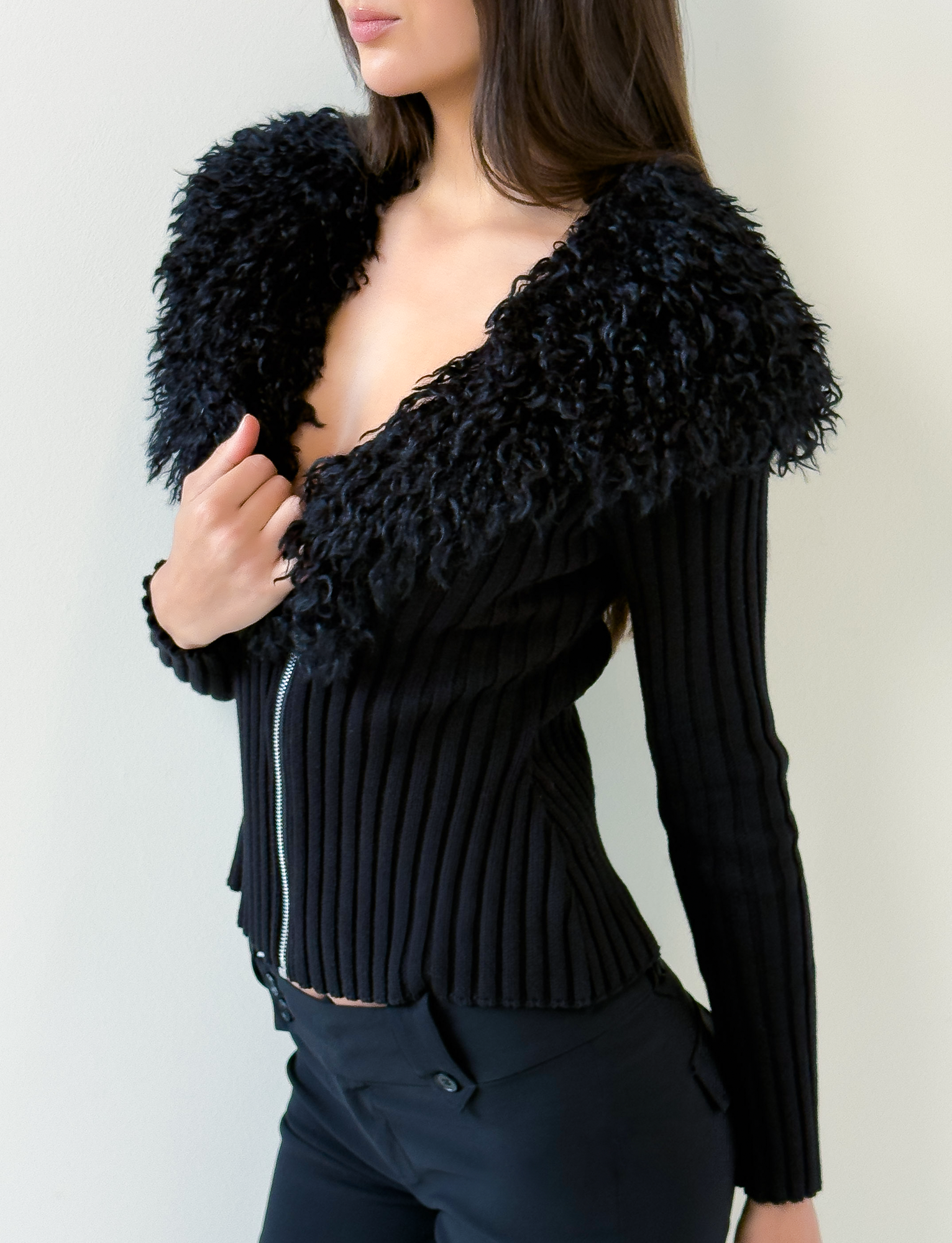 A woman wears the TIGERMIST BRISA JUMPER - BLACK, a fitted knit cardigan with a silver zipper and large faux fur collar, paired with black pants, standing against a plain light wall. Her face is partially out of frame.