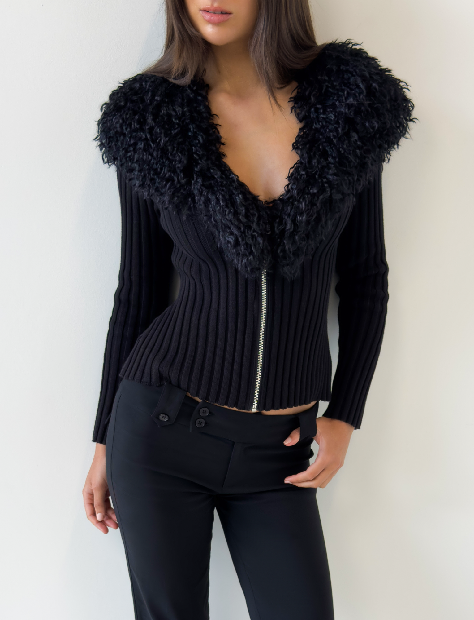 A woman wears the TIGERMIST BRISA JUMPER in black—featuring a ribbed knit design and a large faux fur collar—with black pants, standing against a white wall. Her straight hair is loose, and her face is partly out of frame.