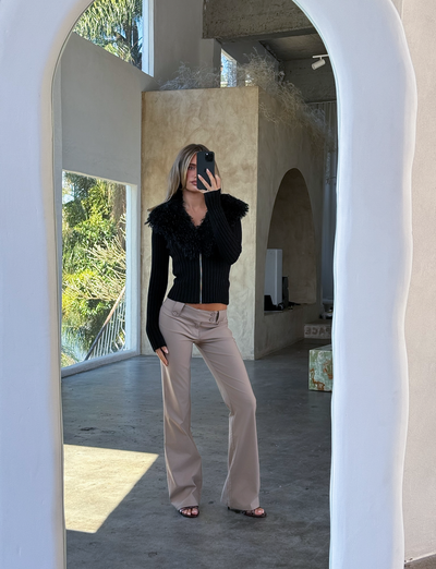 Indoors, a person takes a mirror selfie in the sunlit room wearing the TIGERMIST BRISA JUMPER - BLACK, featuring a ribbed design with faux fur collar, paired with beige pants. Large windows reveal palm trees and stylish arched doorways.