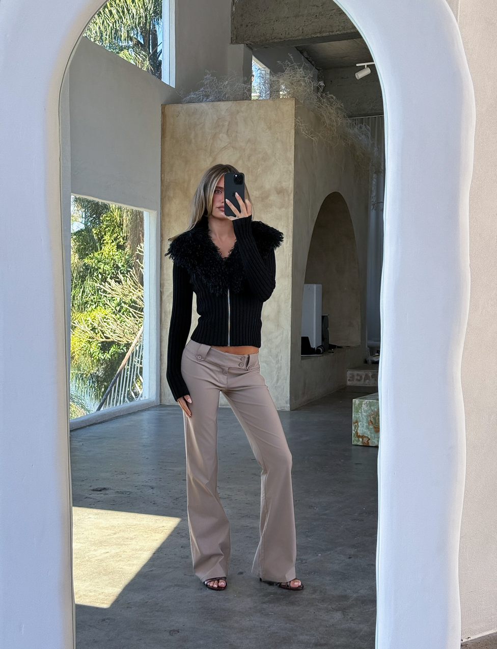 Indoors, a person takes a mirror selfie in the sunlit room wearing the TIGERMIST BRISA JUMPER - BLACK, featuring a ribbed design with faux fur collar, paired with beige pants. Large windows reveal palm trees and stylish arched doorways.
