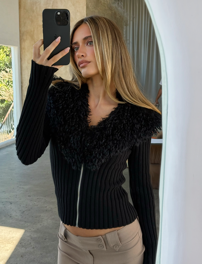 A woman with long blonde hair takes a mirror selfie in a bright room, wearing the TIGERMIST BRISA JUMPER - BLACK paired with beige pants.