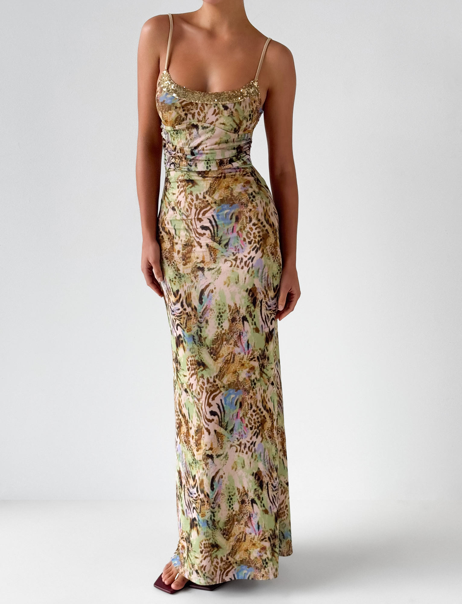 A person stands against a white background wearing the TIGERMIST ELYSIUM MAXI DRESS - ANIMAL SWIRL, featuring a yellow animal print, thin straps, and a sequin neckline.