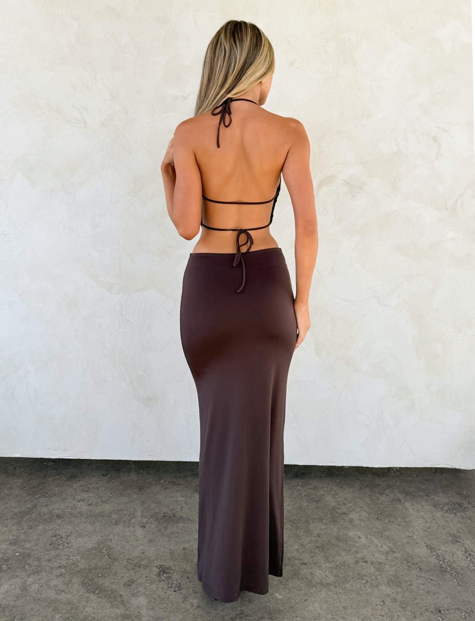A woman faces away wearing the TIGERMIST SURIYA MAXI DRESS in chocolate brown with adjustable halter straps and a gold waist chain, set against a light textured wall and concrete floor.
