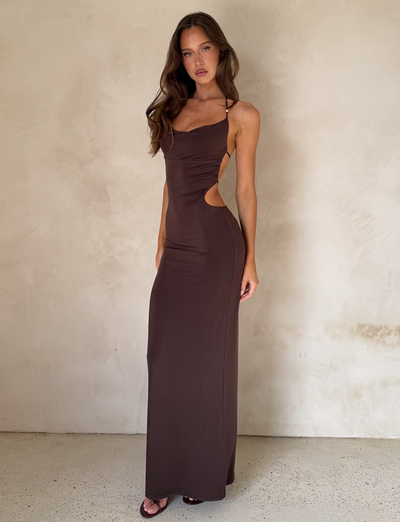A woman wears the TIGERMIST SURIYA MAXI DRESS in chocolate brown, featuring a cowl neck and side cutouts. She pairs it with black open-toe heels, a gold waist chain, and lets her long wavy brown hair frame her look.