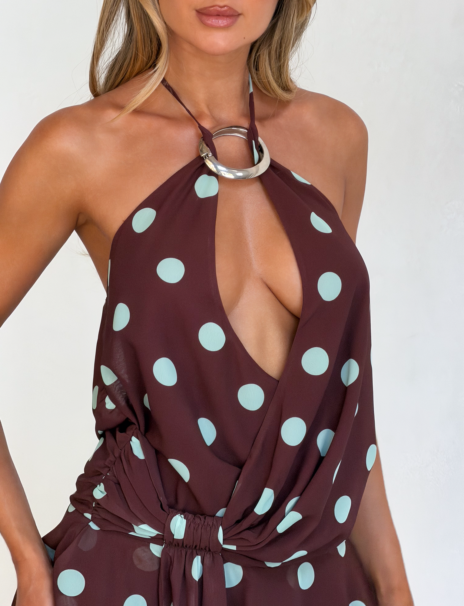 A woman wears the TIGERMIST INDIGO MINI DRESS 2.0 in Chocolate Blue Dot, a sleeveless halter style with large light blue polka dots, a metal ring neckline, deep V-cut, and gathered waist revealing her chest and midriff.