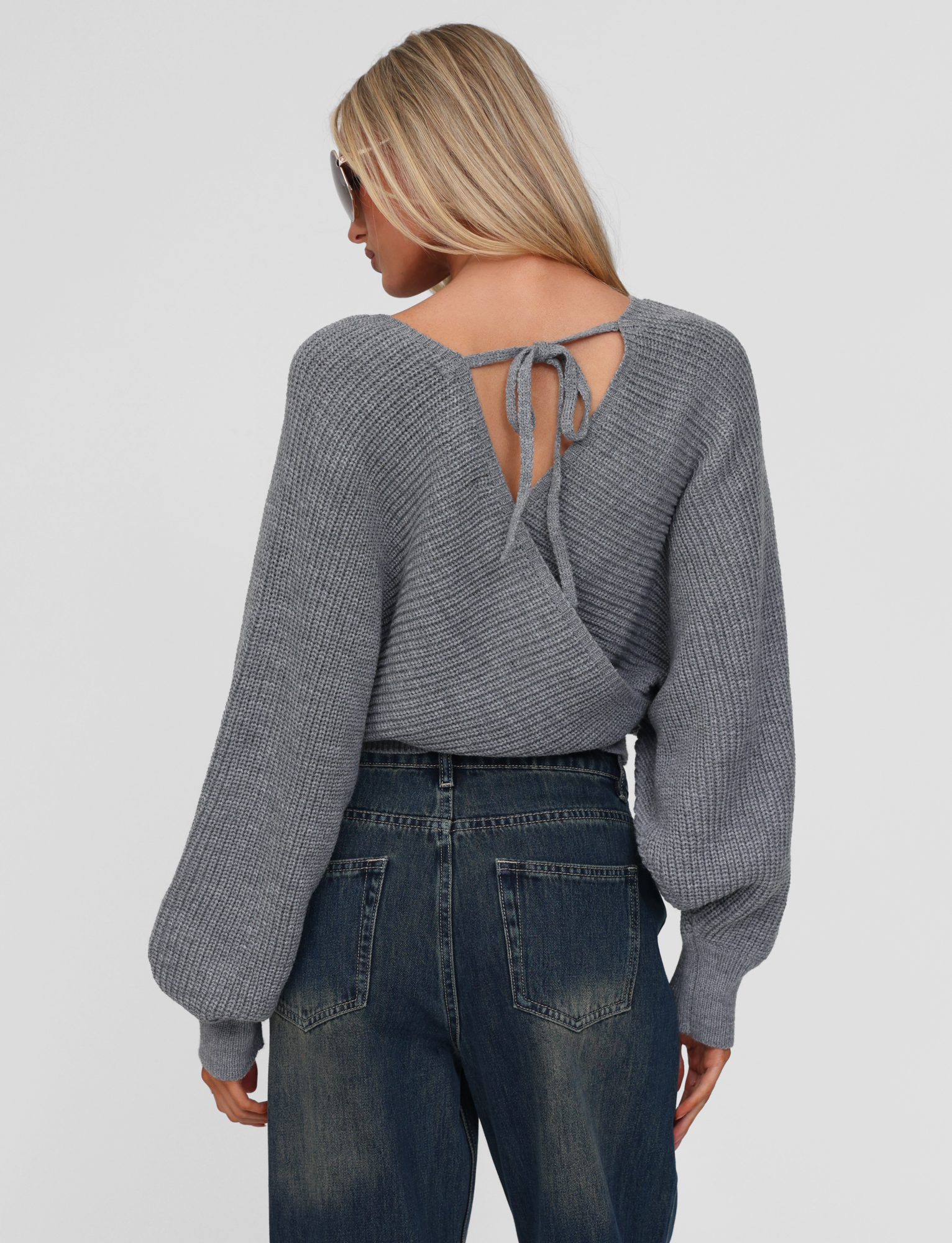 A woman with long blonde hair wears sunglasses, high-waisted blue jeans, and the TIGERMIST RAVEN JUMPER in grey featuring a crisscross back and tie detail. She faces away from the camera against a plain background.