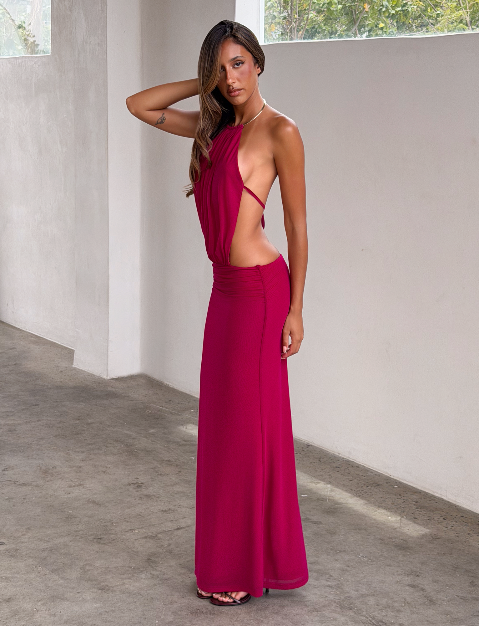 A woman poses indoors in the TIGERMIST EVANNA MAXI DRESS - BERRY, a sleeveless hot pink gown featuring a gold trim neckline, side cutouts, open back, and high slit. She touches her head against a minimalist backdrop of white walls and concrete floor.