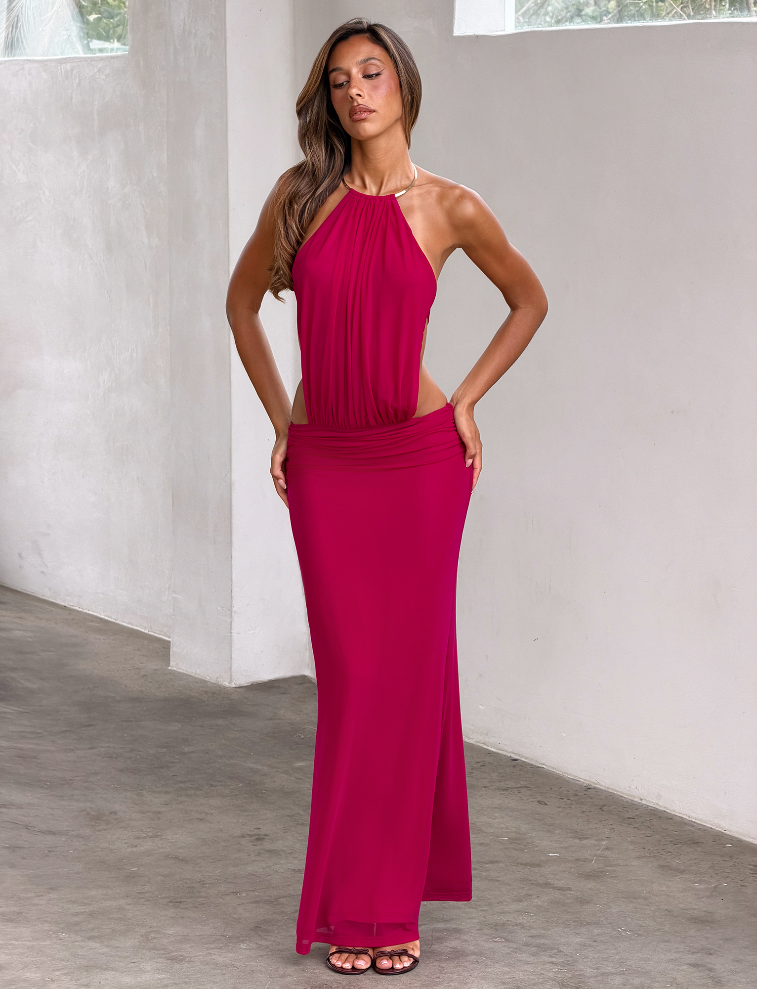 A woman poses confidently indoors wearing the TIGERMIST EVANNA MAXI DRESS in berry, a sleeveless mesh halter style with a gold trim neckline. She stands with hands on her hips against a plain, light background.