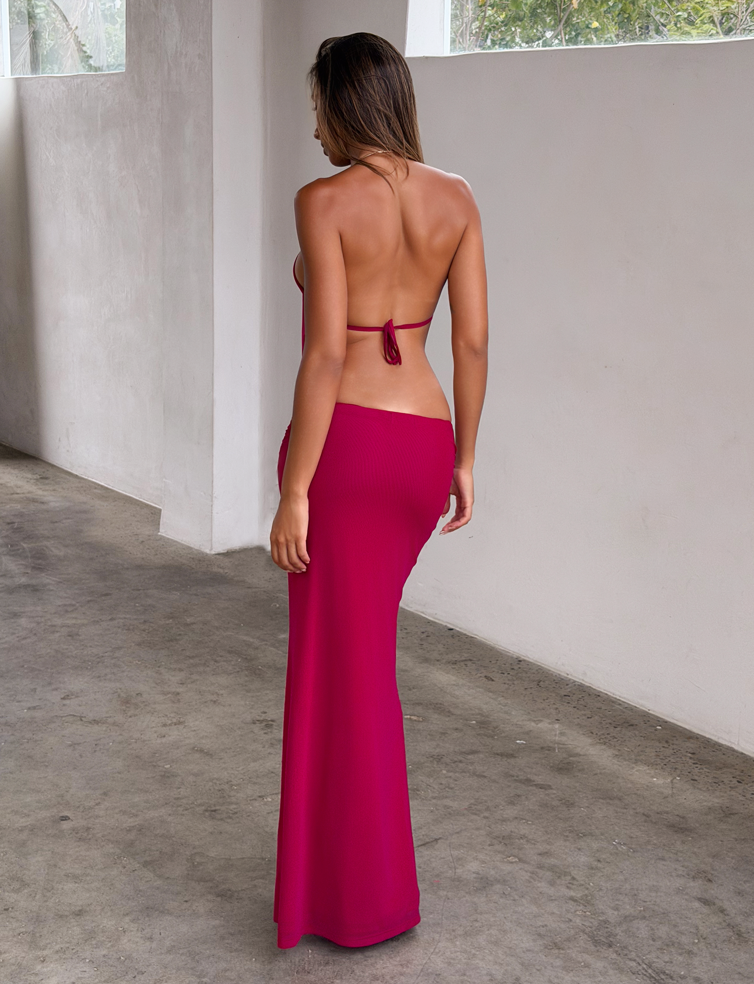 A woman faces away from the camera in a minimalist concrete hallway, wearing the TIGERMIST EVANNA MAXI DRESS in Berry—a bright pink, open-back halter-neck style.