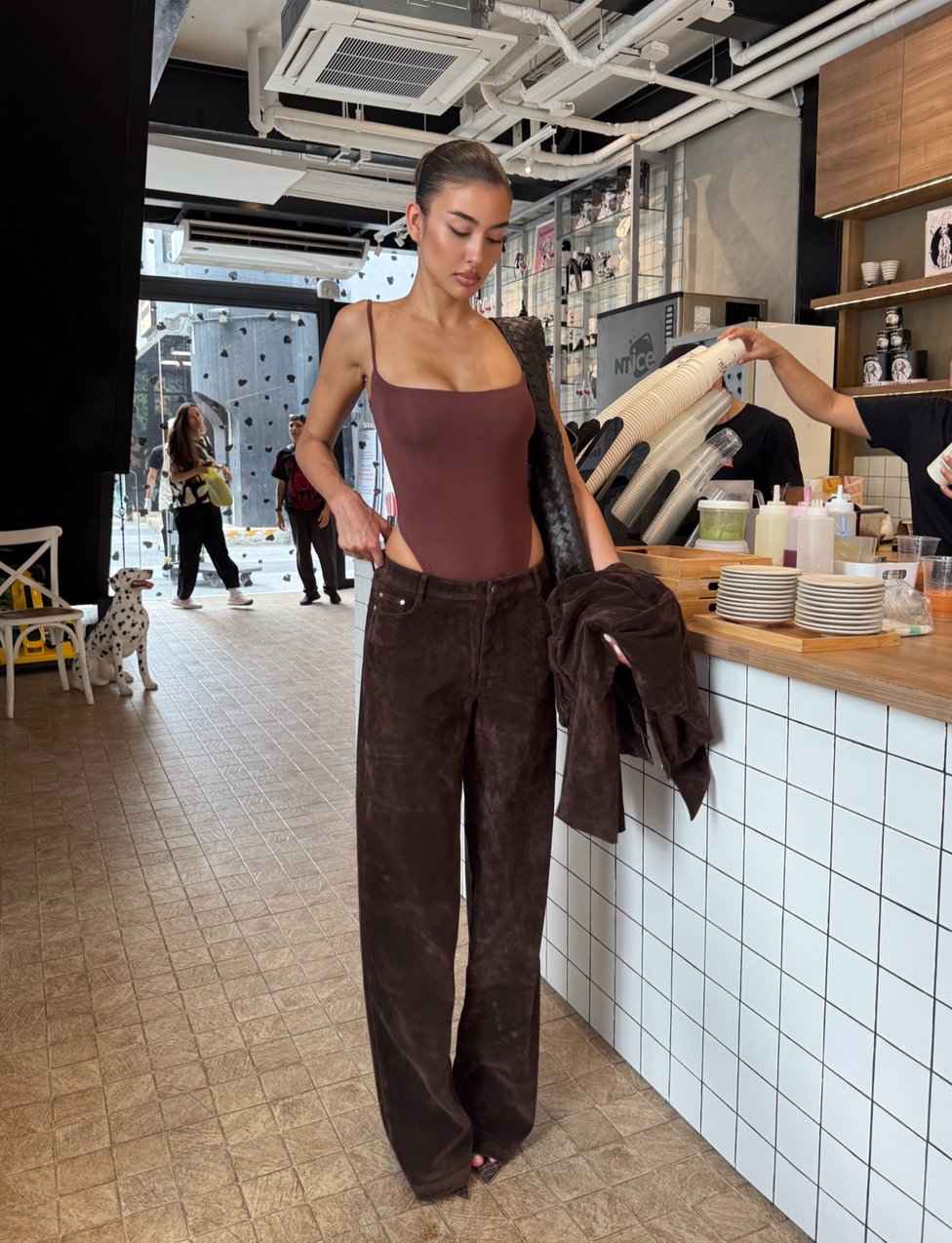 A woman in TIGERMIST's CAMELLIA PANT - CHOCOLATE SUEDE stands at a café counter, holding a matching jacket. The café has a tiled counter, modern decor, and people in the background, including someone with a Dalmatian.
