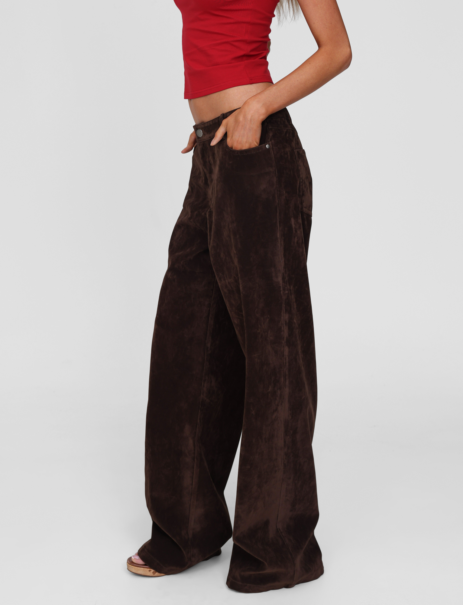 A person stands sideways with one hand in a pocket, wearing the TIGERMIST CAMELLIA PANT in chocolate suede, styled with a red crop top, against a plain white background.