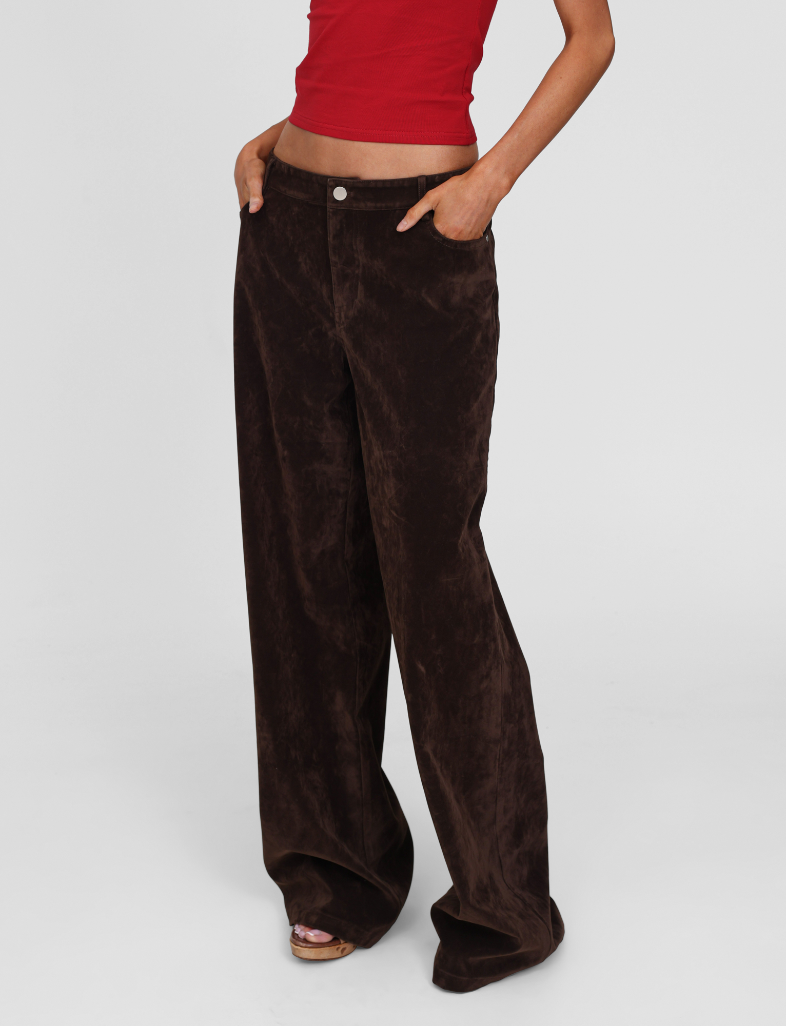 Model wears the TIGERMIST CAMELLIA PANT in chocolate suede, a vintage-washed style, paired with a red sleeveless crop top. Standing hands in pockets against a plain white background. Head not visible in the image.