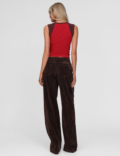 A woman with long blonde hair wears sunglasses and the TIGERMIST CAMELLIA PANT - CHOCOLATE SUEDE, paired with a sleeveless red and brown top, standing facing away from the camera against a plain light background.