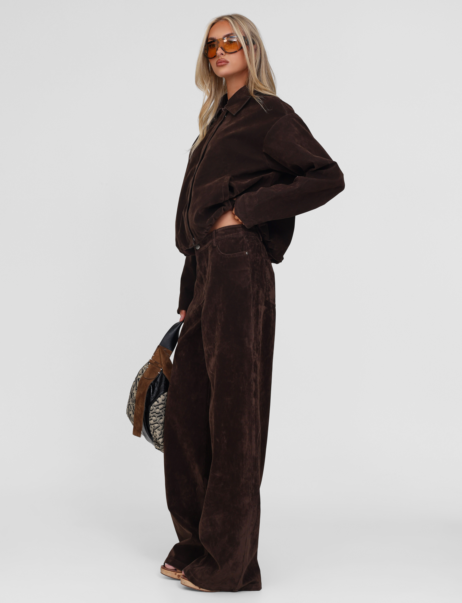 A person stands against a plain white background, wearing the TIGERMIST CAMELLIA PANT in chocolate suede with an oversized dark brown corduroy shirt, sunglasses, and holding a patterned bag.