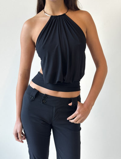 Model wears the TIGERMIST EVANNA TOP - BLACK, a sleeveless halter style with an open back, paired with high-waisted black pants. She stands against a plain white background, one hand on her hip, showcasing the top’s modern elegance.