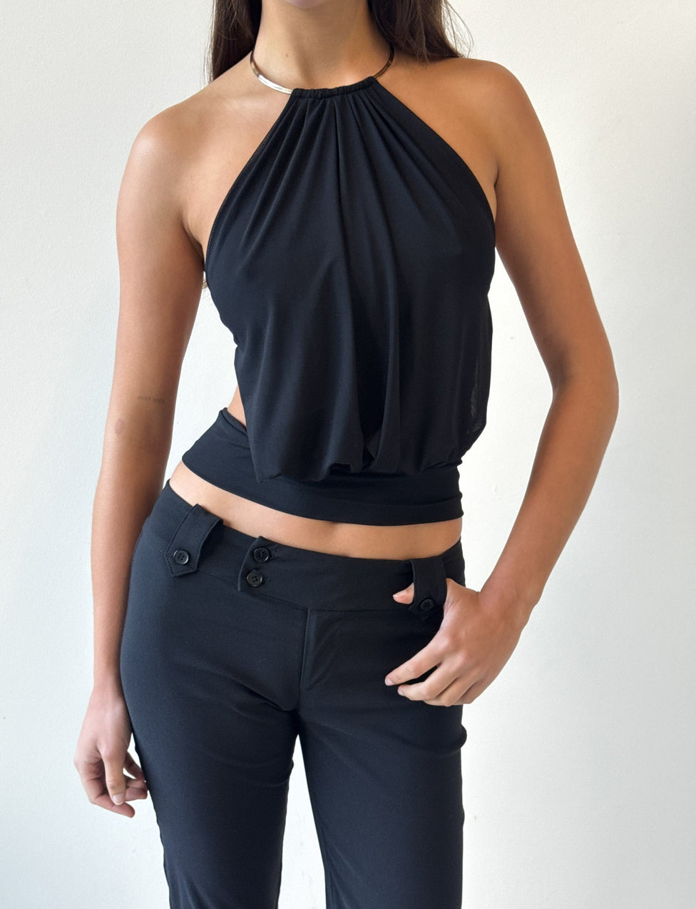 Model wears the TIGERMIST EVANNA TOP - BLACK, a sleeveless halter style with an open back, paired with high-waisted black pants. She stands against a plain white background, one hand on her hip, showcasing the top’s modern elegance.