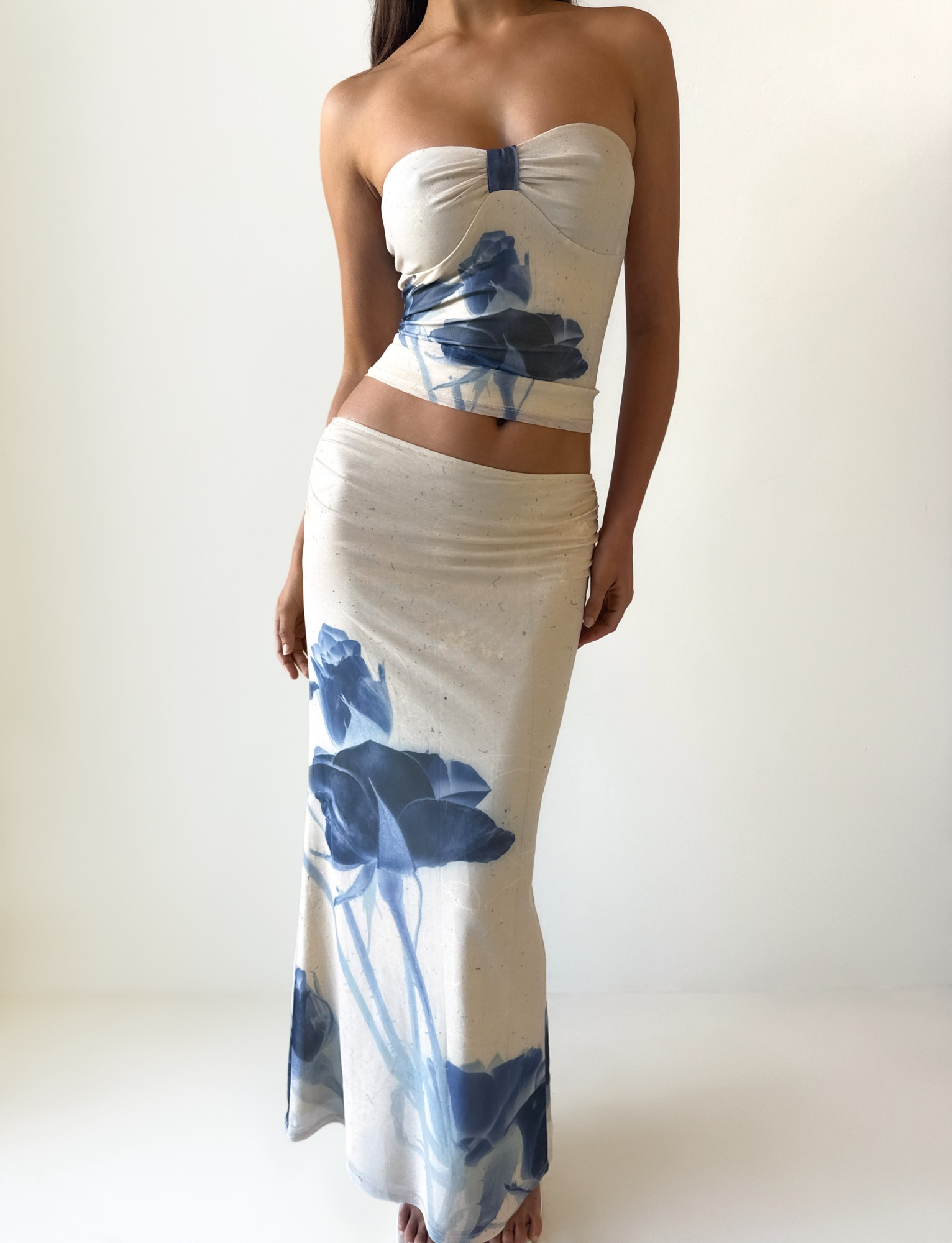 A woman wears the TIGERMIST JANIS TOP - INDIGO FLORAL, a strapless sweetheart tube top in beige mesh with blue floral print, paired with a matching long skirt, standing against a plain white background.