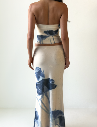 A woman faces away, wearing the TIGERMIST JANIS MAXI SKIRT in indigo floral—a floor-length cream skirt with large blue flower prints—against a plain white background.
