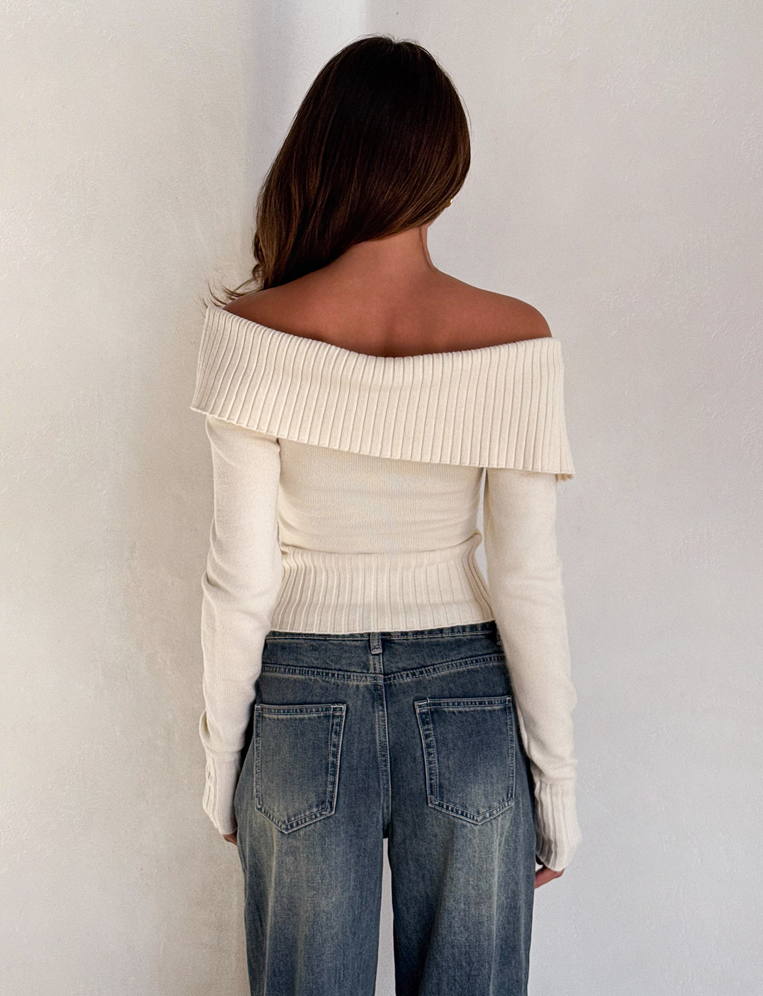 A woman faces away, wearing the TIGERMIST KIERA JUMPER in cream paired with high-waisted blue jeans against a plain light-colored wall.