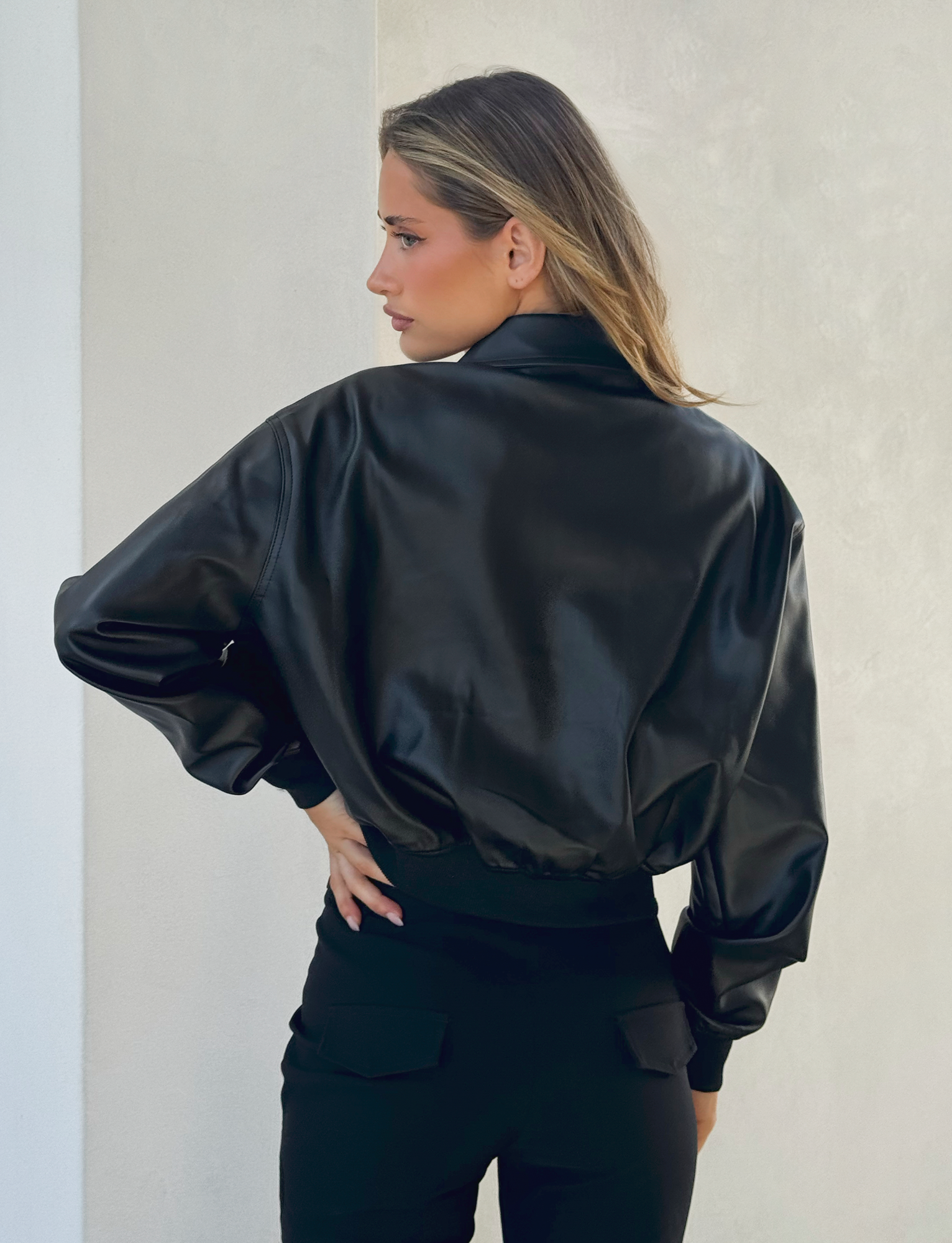 A woman with long hair stands with her back to the camera, wearing the TIGERMIST BEREN JACKET in black and matching pants, in front of a light-colored wall.