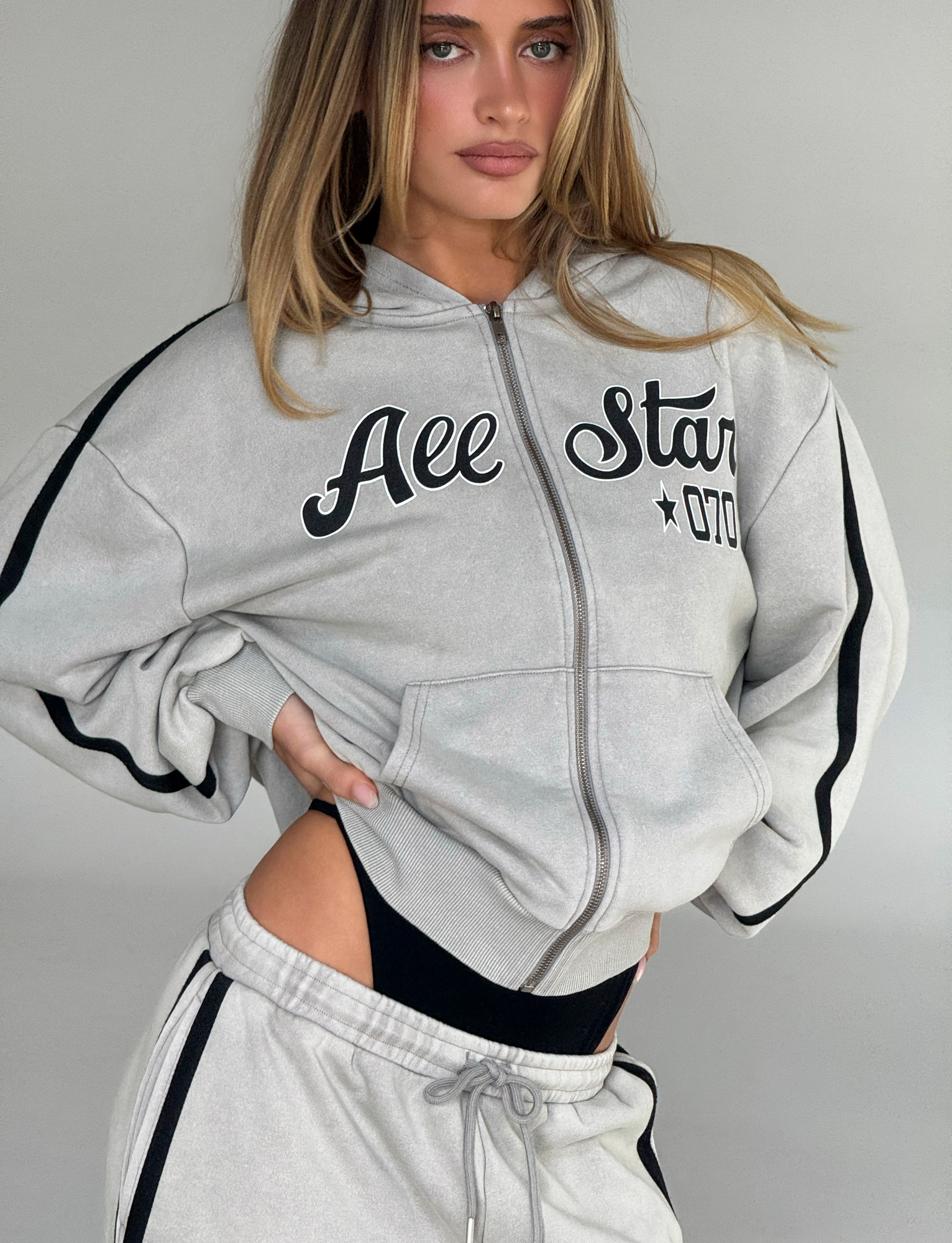 A woman poses with one hand on her hip wearing a vintage-inspired, stone PAYTON HOODIE by TIGERMIST and matching sweatpants, black underwear visible above the waistband, set against a plain background.