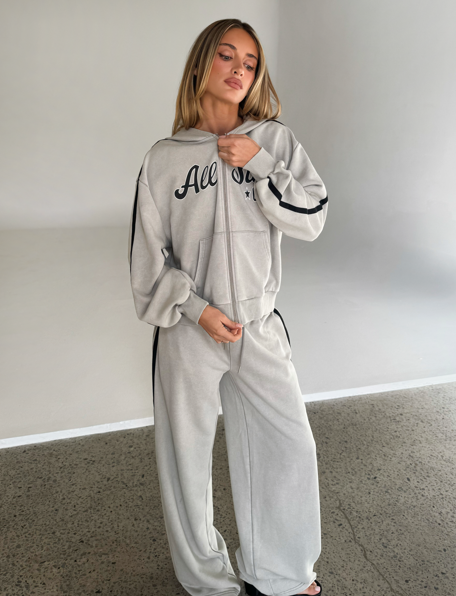 A woman with straight blond hair stands indoors on a polished concrete floor, wearing the TIGERMIST PAYTON HOODIE in Stone and a matching tracksuit. She looks slightly to the side.