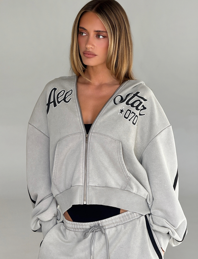 A woman with straight, light brown hair wears the TIGERMIST PAYTON HOODIE in stone, a vintage-inspired, 100% cotton zip-up with matching drawstring pants. She stands against a plain gray background, looking slightly to the side.