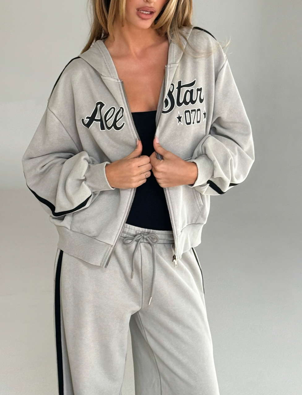 A woman wears the TIGERMIST PAYTON HOODIE in stone with "All Star 07D" on the front and matching sweatpants. She holds the lightweight terry hoodie near the zipper, revealing a black top underneath, against a plain neutral background.