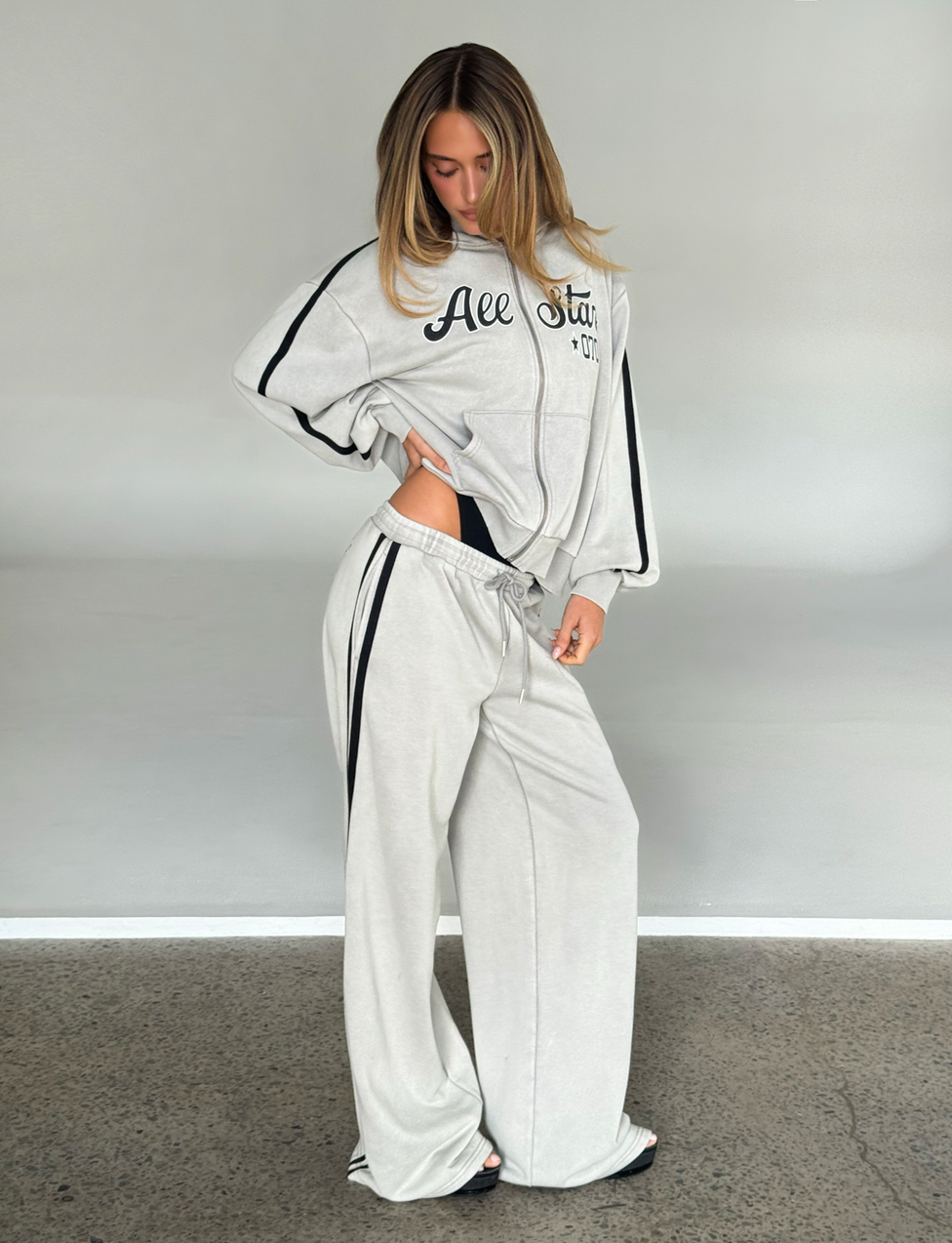 A woman stands indoors wearing the TIGERMIST PAYTON TRACKPANT in stone, a loose-fitting light gray trackpant with black stripes, looking down and slightly tucking her hand into its elastic waistband.