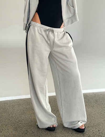A person stands on a speckled floor wearing TIGERMIST PAYTON TRACKPANT - STONE, featuring a light gray tone with a black side stripe, paired with a black bodysuit and platform sandals. The upper body is cropped out of the frame.