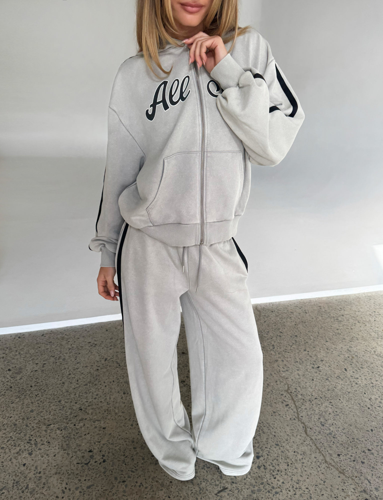 A woman wears TIGERMIST's PAYTON TRACKPANT in stone and a matching light gray tracksuit with black stripes and "All" on the hoodie, standing on a polished concrete floor against a white background; her face is partially out of frame.