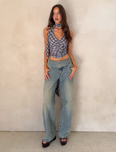 A woman with long, wavy hair and a neutral expression stands against a light wall wearing TIGERMIST's REEF JEAN - LIGHTWASH with detachable belt, paired with a sleeveless plaid crop top, matching scarf, and platform sandals.