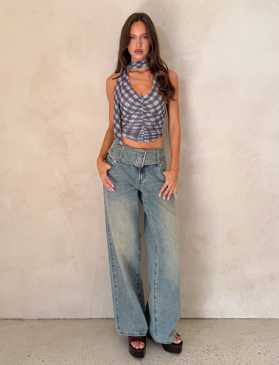 A woman with long brown hair and a neutral expression wears TIGERMIST’s REEF JEAN - LIGHTWASH, styled with a blue plaid halter crop top and black platform sandals, standing against a plain beige wall.